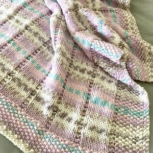 May include: A hand-knitted blanket featuring a textured pattern with alternating stripes of soft pink, light blue, cream, and beige yarn. The knit construction displays a mix of garter and stockinette stitches, creating a varied surface texture throughout.