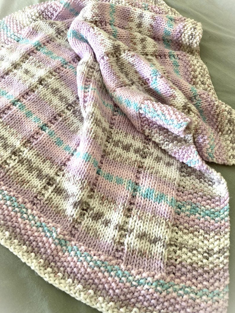 May include: A hand-knitted blanket featuring a textured pattern with alternating stripes of soft pink, light blue, cream, and beige yarn. The knit construction displays a mix of garter and stockinette stitches, creating a varied surface texture throughout.