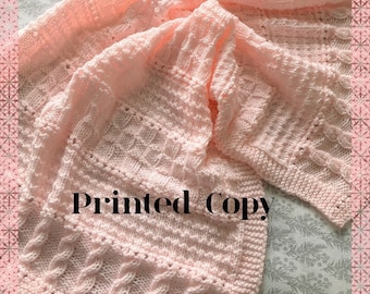 May include: A pink knitted baby blanket with a cable stitch pattern. The blanket is folded and the text "Printed Copy" is visible in the center of the image.