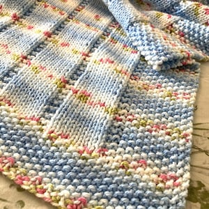 May include: A hand-knitted blanket featuring a textured stitch pattern with alternating vertical stripes of solid light blue and a variegated yarn blend of white, light blue, soft pink, and pale green colours throughout the entire fabric.