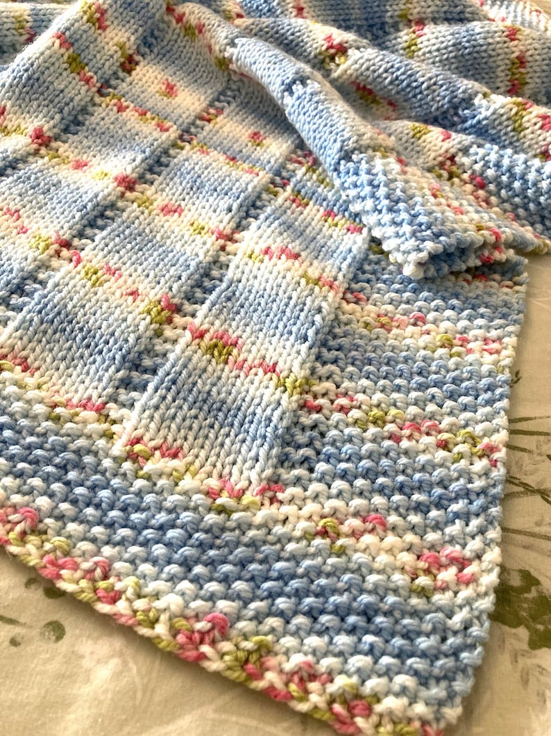 May include: A hand-knitted blanket featuring a textured stitch pattern with alternating vertical stripes of solid light blue and a variegated yarn blend of white, light blue, soft pink, and pale green colours throughout the entire fabric.