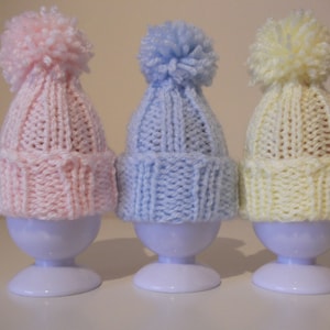 May include: Three pastel-colored, knitted baby hats with pom-poms atop white egg cups. The hats are pink, blue, and yellow. The hats are designed to fit over the egg cups.
