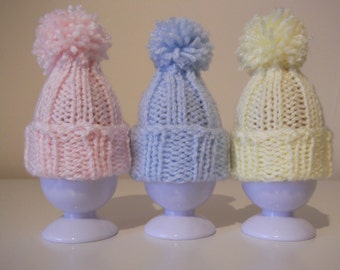 May include: Three pastel-colored, knitted baby hats with pom-poms atop white egg cups. The hats are pink, blue, and yellow. The hats are designed to fit over the egg cups.