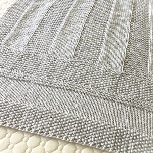 May include: A gray knitted baby blanket with a textured pattern. The blanket is made with a soft yarn and has a simple, modern design.