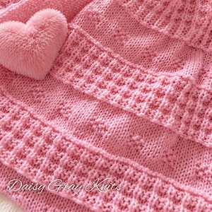 May include: A light pink knitted blanket featuring a textured waffle stitch pattern and alternating rows of small raised bobbles. A soft, plush, heart-shaped accent in the same shade of pink is attached to the corner. The text Daisy Gray Knits is visible.