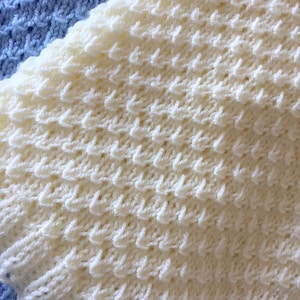 May include: A close-up of a white knitted blanket with a textured pattern. The blanket is made of soft yarn and has a delicate, lacy look.