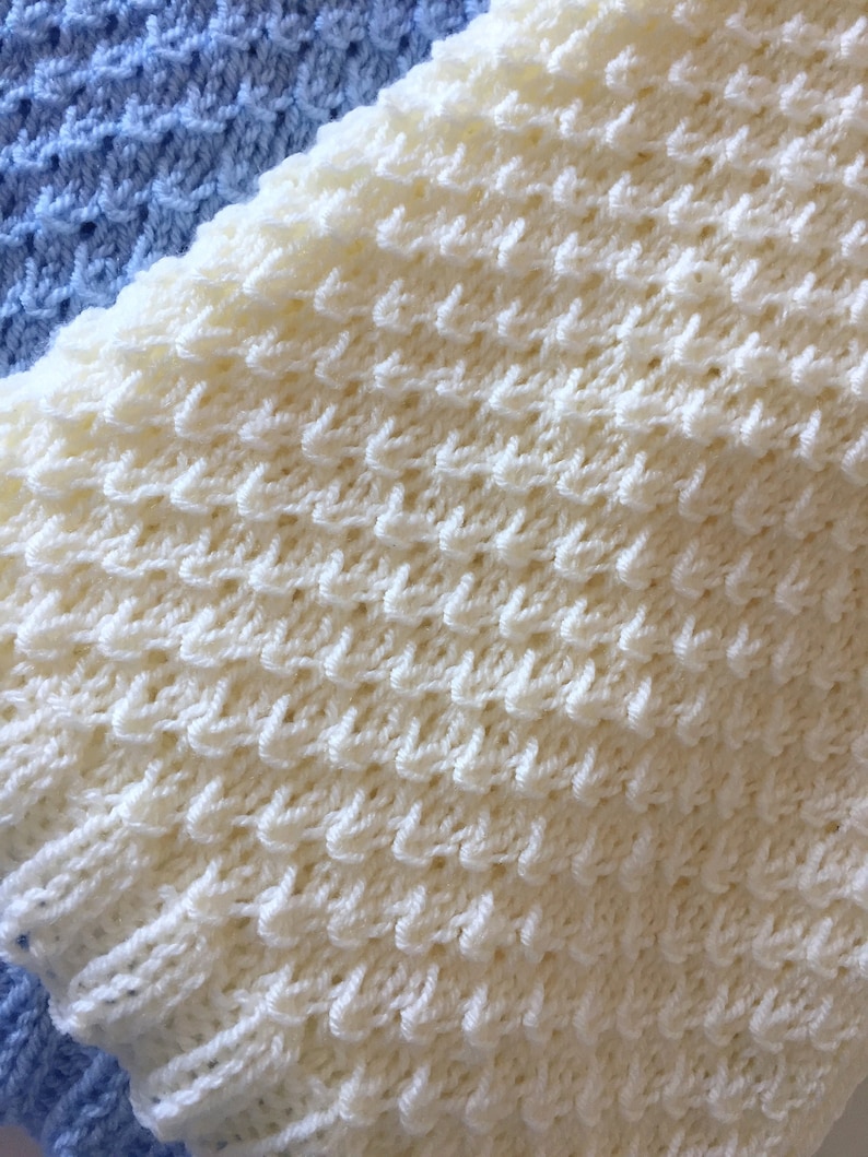 May include: A close-up of a white knitted blanket with a textured pattern. The blanket is made of soft yarn and has a delicate, lacy look.