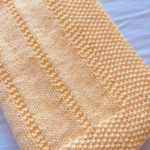 May include: A soft, pale yellow knitted blanket featuring a textured garter stitch pattern. The design includes vertical columns of alternating knit and purl stitches, creating a subtle ribbed effect across the surface of the rectangular textile.