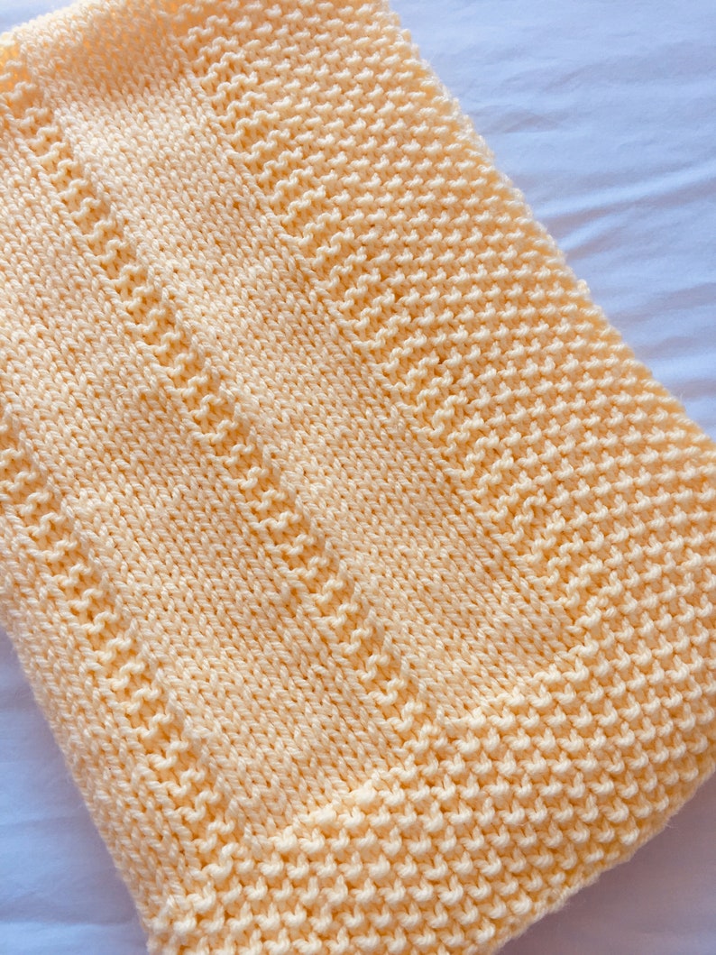 May include: A soft, pale yellow knitted blanket featuring a textured garter stitch pattern. The design includes vertical columns of alternating knit and purl stitches, creating a subtle ribbed effect across the surface of the rectangular textile.