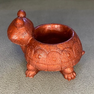May include: A small, metallic copper-coloured turtle-shaped planter with a textured, granular surface. The turtle has a rounded shell that forms a hollow container, a raised head with two small bumps on top, and four short, sturdy legs.
