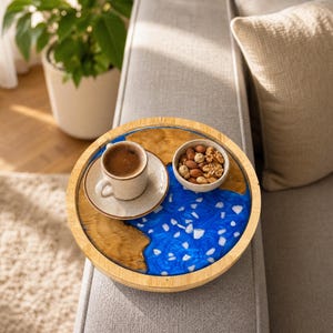 May include: A round wooden serving tray featuring a natural wood grain border and a central inlay of vibrant blue resin with embedded white stone fragments. A small ceramic cup of coffee on a saucer and a bowl of mixed nuts sit on the tray surface.