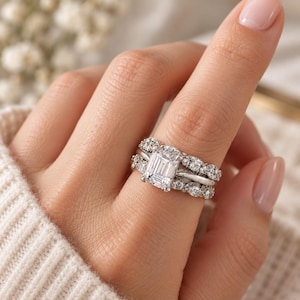 May include: A silver-toned ring set featuring a central emerald-cut stone flanked by smaller round stones. The set includes a main band with a prominent rectangular center stone and a contoured wedding band adorned with multiple small round stones.