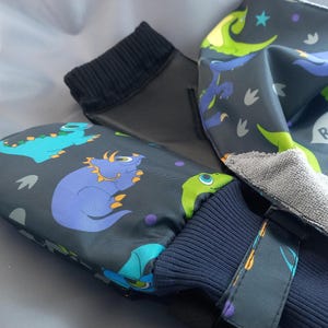 Puede incluir: Un par de manoplas gris oscuro y un babero a juego con un colorido estampado de dinosaurios azules, verdes y morados, estrellas y huellas. El babero incluye el texto ROAR! y DINO, con un reverso de tela gris de textura rugosa.