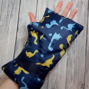 Pode incluir: Uma luva sem dedos azul-escura com um padrão repetitivo de dinossauros de desenho animado em azul-claro e amarelo. A luva tem uma faixa de tecido azul-escuro sólida na zona do pulso e costuras visíveis ao longo das bordas.