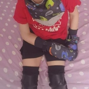 Puede incluir: Un niño con una camiseta roja con el texto CREW, un babero tipo bandana gris oscuro con estampado de dinosaurios, pantalones cortos negros, muñequeras negras con patrones de dinosaurios y rodilleras negras sobre una sábana lila.