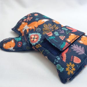 Op de afbeelding: Een donkerblauwe ovenwant met een herhalend patroon van oranje vossen, kleurrijke bloemen en groene bladeren. De want heeft een doorgestikte textuur en een rechthoekige flap aan de achterkant die vastzit met klittenband.