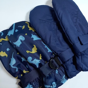 Könnte beinhalten: Ein Paar marineblaue Fäustlinge mit einem sich wiederholenden Muster aus hellblauen und gelben Dinosauriern. Die Fäustlinge verfügen über einen verstellbaren schwarzen Riemen mit Kunststoffschnalle am Handgelenk und Ziernähte.