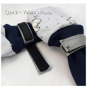 Puede incluir: Un par de manoplas de color gris claro con un gráfico de dinosaurio en líneas negras y puños de punto acanalado azul marino oscuro. Las manoplas incluyen correas de velcro negras ajustables. El texto Type 2 - Velcro strapc es visible.