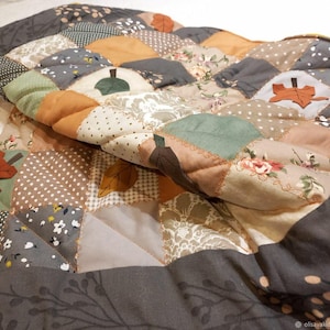 Könnte beinhalten: Eine gesteppte Patchworkdecke mit verschiedenen Stoffquadraten in Grau-, Braun-, Grün- und Cremetönen. Das Design umfasst florale Muster, Punkte und unifarbene Stoffe, akzentuiert durch applizierte Stoffblätter und sichtbare Steppnähte.