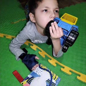 May include: A blue and yellow plastic toy truck with black wheels and a small square fabric book featuring a white patch, a red fabric loop, and a multi-colored geometric pattern with blue, yellow, and pink shapes on a white background.