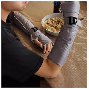 Puede incluir: Un par de mangas protectoras acolchadas de color gris claro con correas negras ajustables y hebillas de plástico. Estas mangas cubren los antebrazos y se extienden hasta las manos, con una abertura para el pulgar. La tela tiene textura.