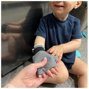 May include: A gray fabric hand mitt with a black hook-and-loop fastener strap around the wrist. The mitt is designed to cover the palm and back of the hand, featuring a smooth, solid gray exterior material and a white interior lining.