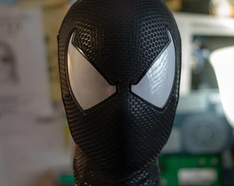 May include: A black full-head mask featuring a textured honeycomb pattern across the entire surface. The mask includes two large, angular, silver-white eye lenses with defined black borders, creating a sleek and uniform appearance.