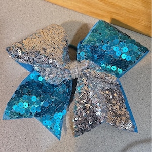 Op de afbeelding: Een grote haarstrik met een gespleten ontwerp, waarbij de ene kant bedekt is met glinsterende zilveren lovertjes en de andere kant met levendige turquoise lovertjes. Het midden is samengebonden met een band van zilveren glitterstof.