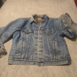 Könnte beinhalten: Eine mittelblaue Jeansjacke mit spitzem Kragen, durchgehender Knopfleiste, zwei Brusttaschen mit geknöpften Klappen und zwei vertikalen Seitentaschen. Die Jacke hat ein kleines rotes Etikett an der linken Brusttasche und ein beiges Label am Nacken.