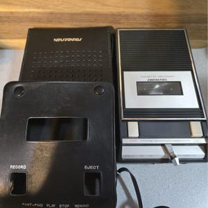 May include: A black Soundesign cassette recorder with a silver control panel and a matching black leather-like carrying case. The recorder features buttons for record, fast-forward, play, stop, rewind, and eject, with text labels printed on both the device and the case.