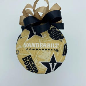 May include: A round, fabric-covered ornament featuring the words "VANDERBILT COMMODORES" and "ANCHOR DOWN" in black text on a gold background. A black and gold ribbon bow is attached to the top. The ornament has a star and "V" logo.