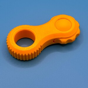 May include: A bright orange plastic fidget tool featuring a circular ring with a textured, gear-like outer edge on one end and a smooth, rounded button-like protrusion on the other end, connected by a solid, slightly contoured central body.