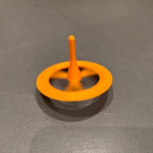 May include: A bright orange spinning top toy featuring a central vertical stem and a circular outer ring. The object is captured in motion, showing a blurred effect around the centre, while the orange plastic structure remains clearly defined.