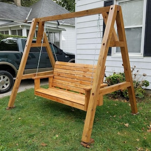 May include: A wooden A-frame swing set featuring a slatted bench seat suspended by metal chains. The structure is constructed from light brown timber beams and includes armrests on both sides of the bench, providing a stable seating area.