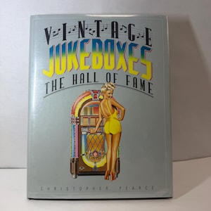 May include: A hardcover book titled Vintage Jukeboxes The Hall of Fame by Christopher Pearce. The light gray cover features a colorful illustration of a classic jukebox and a person in a yellow swimsuit standing beside it, with musical notes above.
