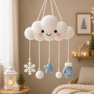 Op de afbeelding: Een wit gehaakte wolkenmobiel met een lachend gezicht en roze wangen. Aan witte koorden met houten kralen hangen een witte sneeuwvlok, twee witte pompons, een lichtblauwe ster en een lichtblauwe want met een sneeuwvlokpatroon eraan vast.