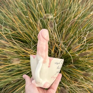 May include: A ceramic mug with a cream-colored base and a pink phallic-shaped handle extending upward. The mug features handwritten cursive text in black ink on the side, with pink glaze dripping down from the handle onto the mug's rim.