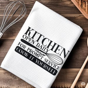 May include: A white waffle-weave kitchen towel featuring black text that reads KITCHEN OPEN DAILY FOR PROMPT SERVICE COOK IT YOURSELF, accompanied by a graphic illustration of a wire whisk centred within the printed text design.