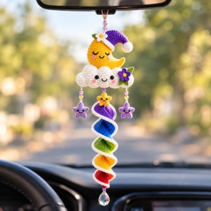 Könnte beinhalten: Ein gehäkelter Auto-Anhänger mit einem gelben Halbmond mit violetter Schlafmütze, einer weißen Wolke mit violetter Blume und einem regenbogenfarbenen Spiralschwanz. Das Design umfasst kleine lächelnde Sterne und ein klares Kristallprisma am unteren Ende.