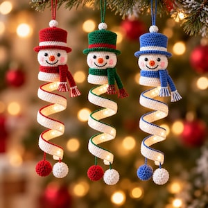 Könnte beinhalten: Drei gehäkelte Schneemann-Ornamente hängen vertikal. Jeder Schneemann trägt einen Zylinder und einen Schal in Rot, Grün oder Blau. Unter jedem Kopf befindet sich ein langer, weißer, spiralförmiger Schwanz mit LED-Lichtern und zwei Bommeln.