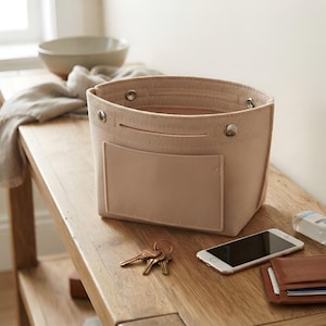 May include: A beige felt storage basket with a rectangular front pocket and silver-toned metal snap buttons along the top rim. The basket features a structured open-top design with a flat base, providing a simple container for organizing small items.