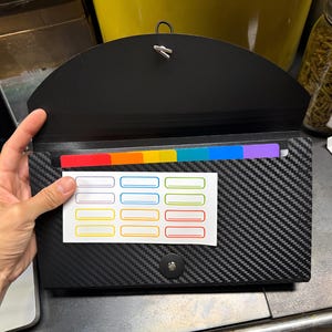May include: A black carbon fiber textured document organizer with a curved flap and a snap closure. The interior features multi-colored dividers in red, orange, yellow, green, blue, and purple. A white sticker sheet with twelve blank rectangular labels is attached.