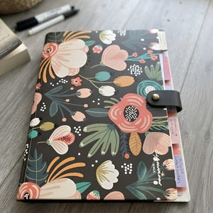 May include: A black planner featuring a colorful floral pattern with pink, peach, teal, and white flowers and leaves. The planner has a black strap closure with a gold-tone snap button and includes several colored index tabs with handwritten text.
