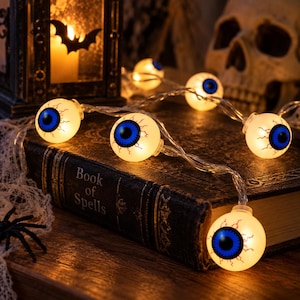 May include: A string of LED lights featuring spherical white eyeballs with bright blue irises and red vein details. The lights are connected by a clear wire and are arranged across a dark, weathered book with the text Book of Spells on the cover.