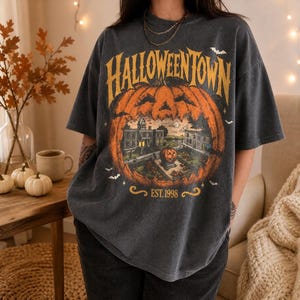 Puede incluir: Una camiseta de manga corta gris carbón con un gráfico grande de una calabaza naranja que contiene una escena urbana. El texto HALLOWEENTOWN está impreso en amarillo sobre la calabaza, con EST. 1998 debajo. Pequeños murciélagos blancos rodean el diseño.