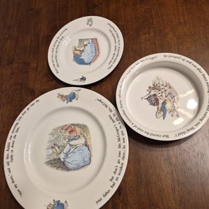 Peut inclure: Trois plats en céramique blanche illustrés avec Peter Rabbit. Une assiette montre Peter au lit, une autre Peter dans un jardin, et un bol montre Peter en train de courir. Chaque pièce comporte du texte imprimé tiré de l'histoire sur le bord.