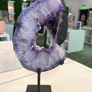 May include: A large, crescent-shaped amethyst geode featuring deep purple crystalline structures on the interior and a dark, rough exterior. The specimen is mounted on a black metal stand with a square base, displaying its natural, jagged edges.