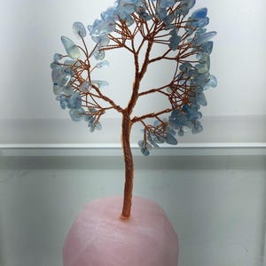 May include: A decorative tree sculpture featuring a twisted copper wire trunk and branches adorned with numerous small, translucent light blue stone chips. The tree is securely anchored into a smooth, rounded base made of solid, pale pink quartz stone.
