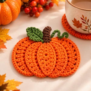 May include: A handmade crochet coaster shaped like a pumpkin, featuring bright orange yarn, a dark brown stem, and a green leaf with a curly vine. The coaster is crafted with visible stitches and a textured surface, designed to hold a beverage mug.