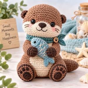 Op de afbeelding: Een gehaakte otterknuffel in bruin en crème die een lichtblauwe gehaakte vis vasthoudt. De otter draagt een blauwgroene sjaal vastgezet met een kleine houten knoop met een pootafdruk. Het heeft zwarte ogen, roze wangen en donkerbruine voetzooltjes.